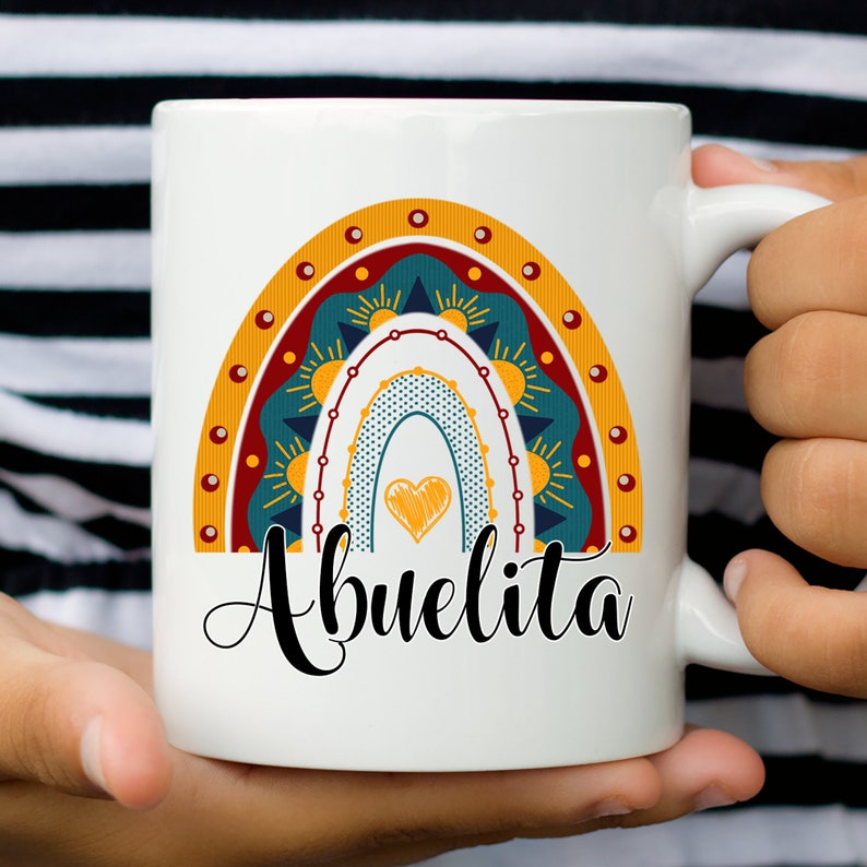 May include: White ceramic mug featuring a colorful rainbow design with a heart and the word "Abuelita" in black script. The rainbow includes shades of teal, orange, and yellow. The mug is held by a person.