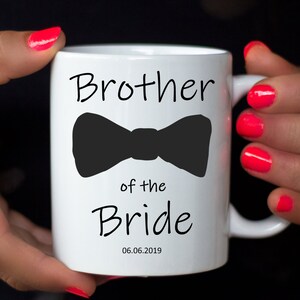 Pu&ograve; includere: Tazza in ceramica bianca con la scritta "Brother of the Bride" e un fiocco nero. La data "06.06.2019" &egrave; stampata sotto. La tazza &egrave; tenuta da una persona con smalto per unghie rosa acceso.