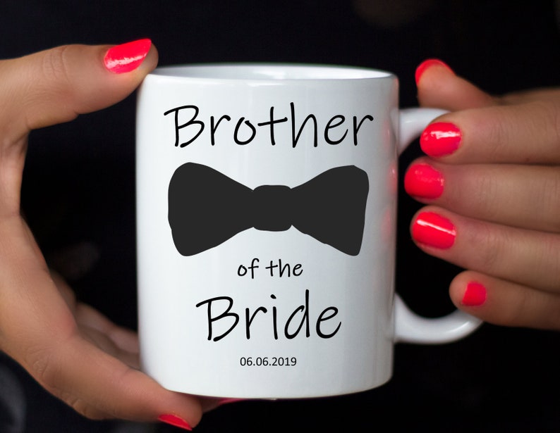 Pu&ograve; includere: Tazza in ceramica bianca con la scritta "Brother of the Bride" e un fiocco nero. La data "06.06.2019" &egrave; stampata sotto. La tazza &egrave; tenuta da una persona con smalto per unghie rosa acceso.