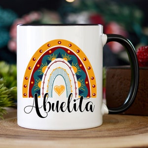 May include: White ceramic mug with a black handle and rim. The mug features a colorful rainbow design with a heart and the word "Abuelita" in black script. The rainbow includes shades of yellow, teal, and red.