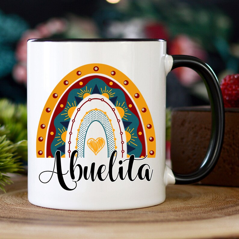 May include: White ceramic mug with a black handle and rim. The mug features a colorful rainbow design with a heart and the word "Abuelita" in black script. The rainbow includes shades of yellow, teal, and red.
