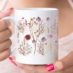 May include: White ceramic mug with a watercolour floral design in shades of brown, purple, and gold. The botanical illustration features various flowers and foliage, creating a natural and elegant aesthetic.