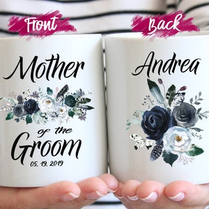 May include: Two white ceramic mugs featuring floral designs in shades of navy blue, grey, and white. The left mug displays the text Mother of the Groom and the date 05.19.2019. The right mug features the name Andrea in black script lettering.