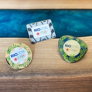 Puede incluir: Tres pines de oro con un diseño en negro, blanco y verde. Cada pin presenta el texto "RIO 2016" y los anillos olímpicos.