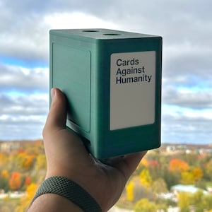 以下が含まれることがあります： 前面に「Cards Against Humanity」と書かれた緑色の箱。箱は人の手に持たれている。
