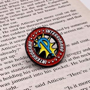 Puede incluir: Un pin de esmalte rojo, blanco, azul y amarillo con un signo de interrogación estilizado y las palabras "Interrobang" escritas dos veces alrededor del borde.