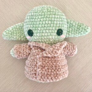 May include: A green and pink crocheted plush toy in the shape of a character from Star Wars. The toy is wearing a pink crocheted coat.