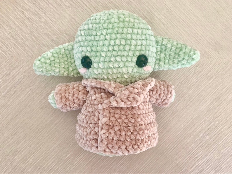 May include: A green and pink crocheted plush toy in the shape of a character from Star Wars. The toy is wearing a pink crocheted coat.