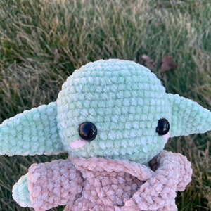 May include: A green crocheted plush toy with large black eyes and a pink blush. The toy is wearing a brown crocheted coat.