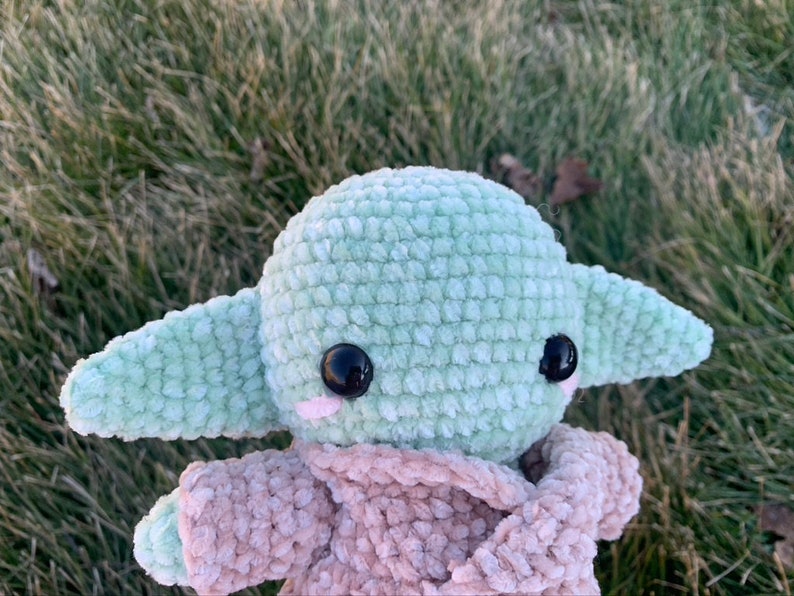 May include: A green crocheted plush toy with large black eyes and a pink blush. The toy is wearing a brown crocheted coat.