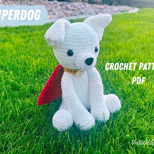 Puede incluir: Un perro blanco tejido a ganchillo con una capa roja, sentado sobre hierba verde. El perro tiene ojos negros y una nariz negra. El texto en la imagen dice "SUPERDOG" y "CROCHET PATTERN PDF".