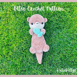 May include: A crocheted otter amigurumi toy holding a blue fish. The otter is brown and has a white face with black eyes. The toy is sitting on a green grassy surface. The text "Otter Crochet Pattern" is at the top of the image and "KatieKittyCraft" is at the bottom of the image.