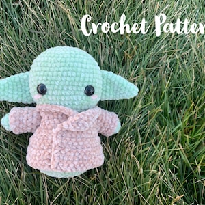 May include: Crochet pattern for a green and pink plush toy resembling a character from the Star Wars franchise. The toy is sitting on a bed of green grass.