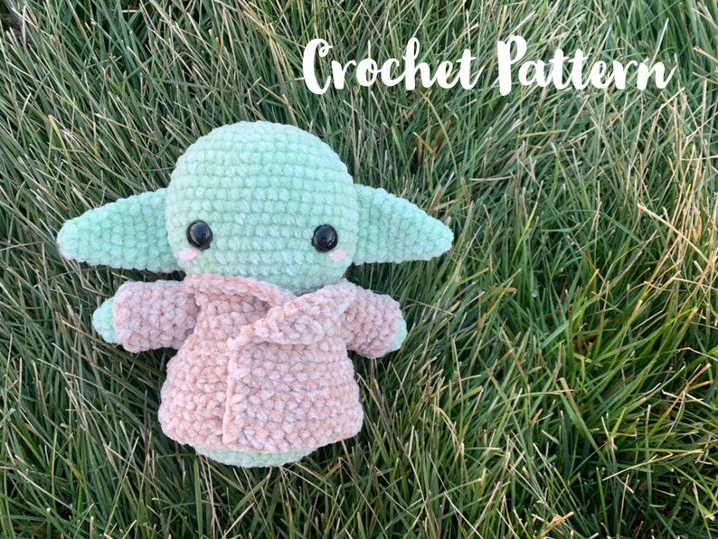 May include: Crochet pattern for a green and pink plush toy resembling a character from the Star Wars franchise. The toy is sitting on a bed of green grass.