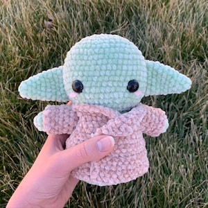May include: A green crocheted plush toy with large eyes and a pink and brown crocheted coat. The toy is being held in a hand.