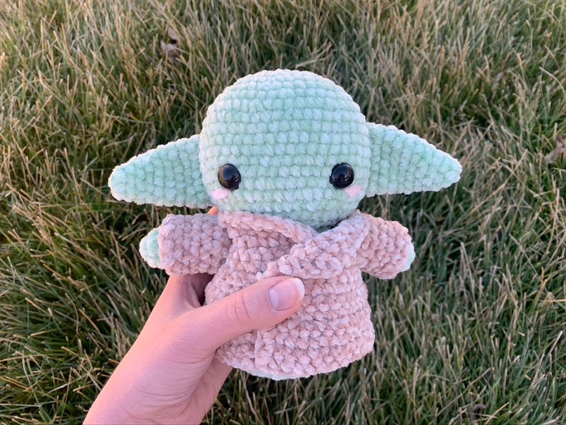 May include: A green crocheted plush toy with large eyes and a pink and brown crocheted coat. The toy is being held in a hand.