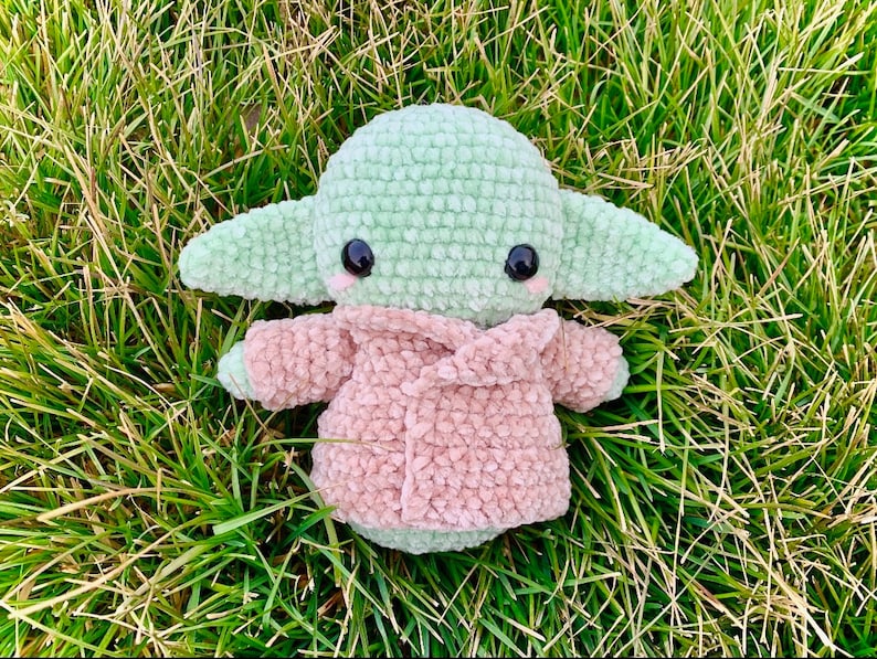 May include: A green crocheted plush toy with large eyes and a pink crocheted jacket. The toy is sitting on a bed of green grass.