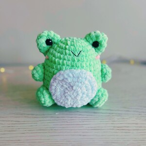 Puede incluir: Un juguete de peluche de rana verde tejido a crochet con una barriga peluda azul claro. La rana tiene ojos negros y una nariz rosa.