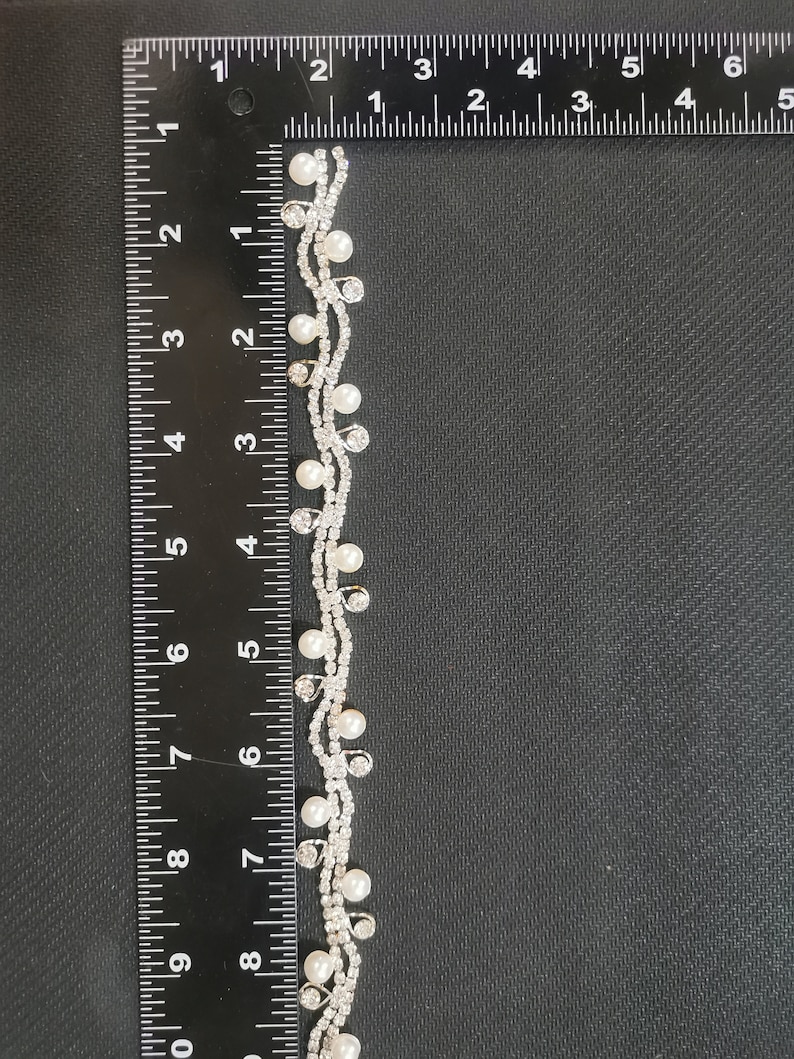 May include: A silver and white beaded trim with small pearls and rhinestones. The trim is measured against a ruler, showing it is approximately 7 inches long.