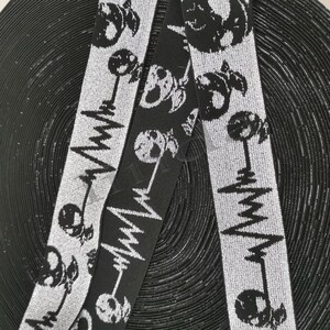 Könnte beinhalten: Nahaufnahme von zwei Rollen elastischem Band mit einem sich wiederholenden Muster aus Totenköpfen und Herzfrequenzmonitorlinien. Ein Band ist schwarz mit weißem Design, das andere ist silber mit schwarzem Design. Die Bänder befinden sich auf schwarzem Hintergrund.