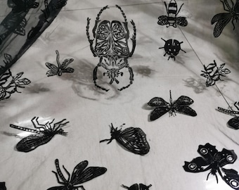 Peut inclure: Tissu noir orné d'illustrations d'insectes, notamment des scarabées, des papillons et des libellules. Les dessins sont détaillés et variés. Le tissu est semi-transparent, avec des motifs d'insectes plus foncés, créant un contraste.