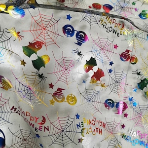 K&ouml;nnte beinhalten: Wei&szlig;er Stoff mit einem sich wiederholenden Muster aus bunten, schillernden Halloween-Motiven. Die Motive umfassen Spinnen, Flederm&auml;use, Geister, K&uuml;rbisse, Sterne und die Worte "Happy Halloween".