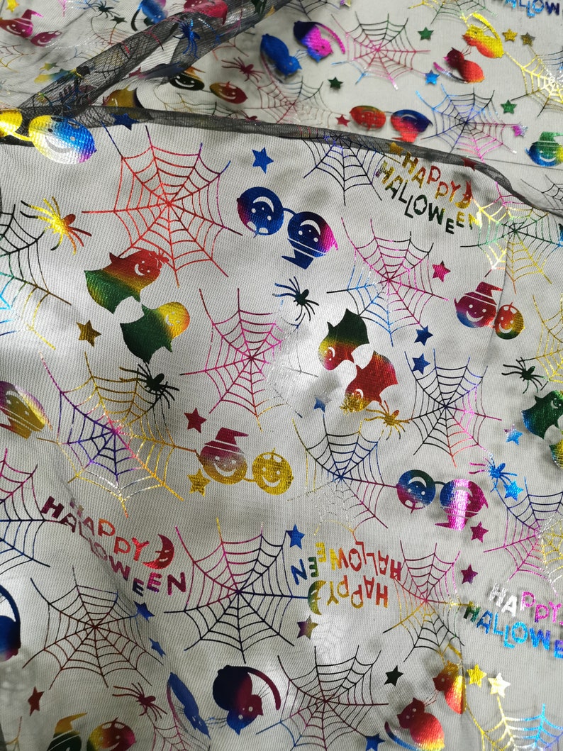 K&ouml;nnte beinhalten: Wei&szlig;er Stoff mit einem sich wiederholenden Muster aus bunten, schillernden Halloween-Motiven. Die Motive umfassen Spinnen, Flederm&auml;use, Geister, K&uuml;rbisse, Sterne und die Worte "Happy Halloween".
