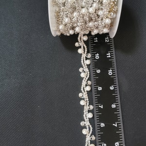 May include: A spool of white pearl and rhinestone trim with a dangling strand of pearls and rhinestones. The trim is approximately 7 inches long.