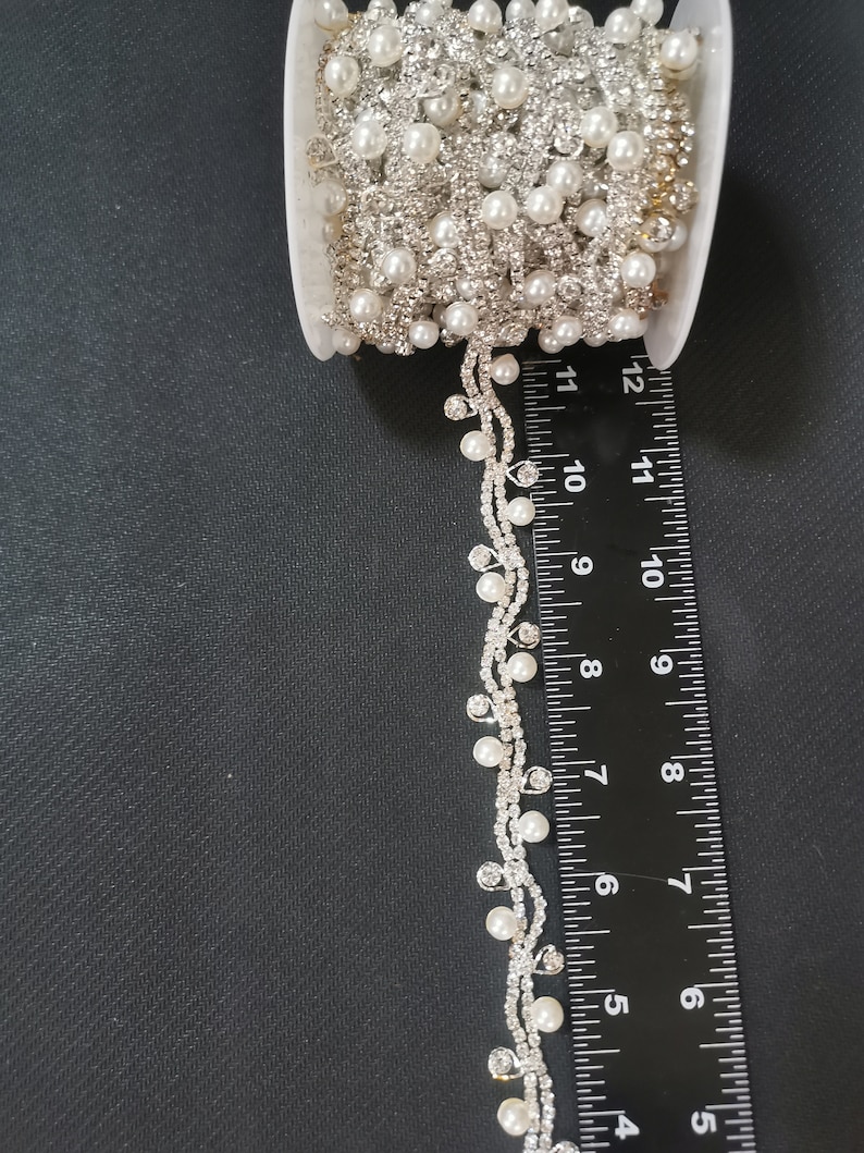 May include: A spool of white pearl and rhinestone trim with a dangling strand of pearls and rhinestones. The trim is approximately 7 inches long.
