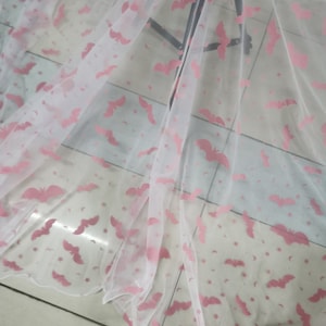 Op de afbeelding: Witte, doorschijnende stof met een roze vleermuis- en sterrenpatroon. De stof is gevouwen en gedrapeerd over een oppervlak.