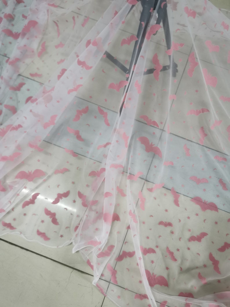 Op de afbeelding: Witte, doorschijnende stof met een roze vleermuis- en sterrenpatroon. De stof is gevouwen en gedrapeerd over een oppervlak.