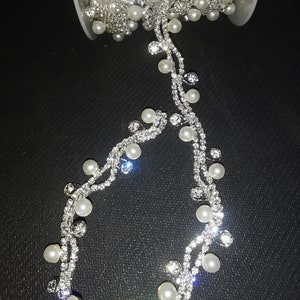 May include: A spool of silver rhinestone trim with white pearl accents. The trim is on a black background.