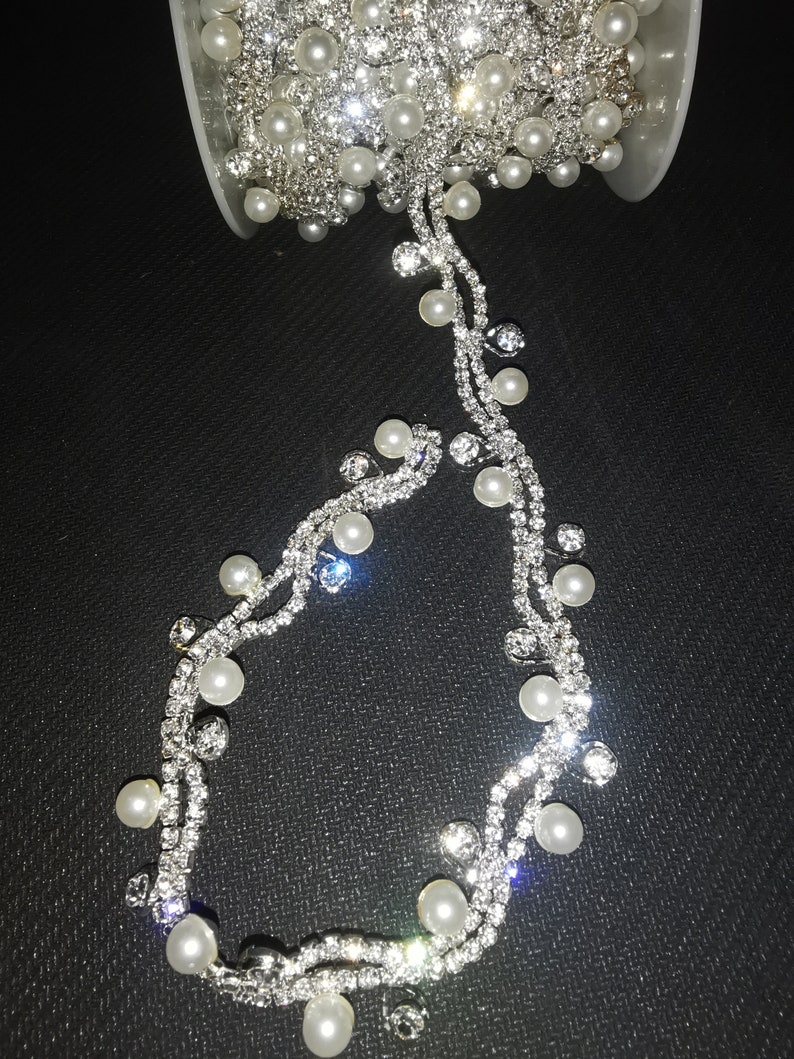 May include: A spool of silver rhinestone trim with white pearl accents. The trim is on a black background.