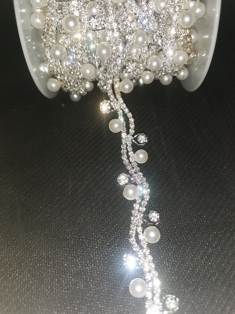 May include: A silver beaded trim with white pearls. The trim is on a spool and a section is hanging down.