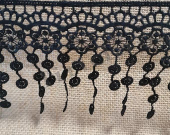 Peut inclure: Garniture en dentelle noire avec un bord festonné et des cercles pendants. La garniture est sur un fond de toile de jute.