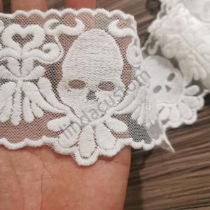 May include: White lace trim featuring a repeating skull design. The lace has a delicate, open weave with detailed skull and floral-like motifs. The trim is ideal for adding a unique touch to apparel or craft projects.