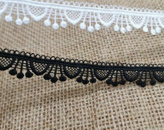 Peut inclure: Gros plan sur deux garnitures de dentelle décoratives. L'une est blanche, l'autre noire. Les deux garnitures présentent un bord festonné avec de petits accents ronds. La dentelle est posée sur un fond texturé beige.