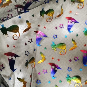 K&ouml;nnte beinhalten: Ein wei&szlig;er Stoff mit einem regenbogenfarbenen, schillernden Muster aus Sternen, Flederm&auml;usen, Katzen und Hexenh&uuml;ten.