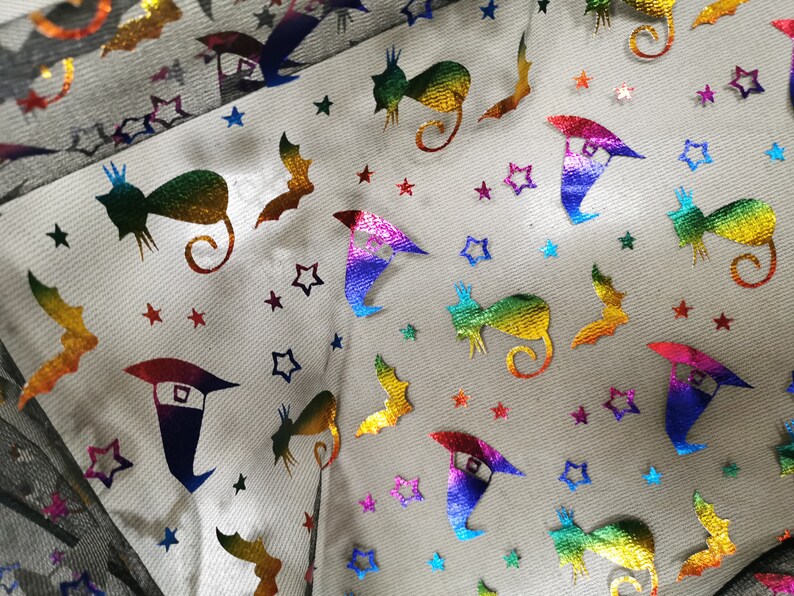 K&ouml;nnte beinhalten: Ein wei&szlig;er Stoff mit einem regenbogenfarbenen, schillernden Muster aus Sternen, Flederm&auml;usen, Katzen und Hexenh&uuml;ten.