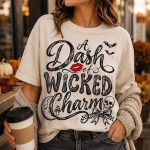 May include: A cream-colored t-shirt featuring black text that reads A Dash of WICKED Charm. The design includes a red lip icon, two small bats, a crescent moon with a face, a small mushroom, a skull, and a bow with a checkered pattern.