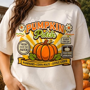 Könnte beinhalten: Ein cremefarbenes T-Shirt mit einer Grafik eines orangefarbenen Kürbisses mit grünen Blättern. Der Text lautet PUMPKIN PATCH, FRESH FROM THE PATCH, FARM FRESH und FOCALLY BROWN AUTUNH BICKED, mit einem Banner für PUMPKINS, CORN MAZE, APPLE CIDER.