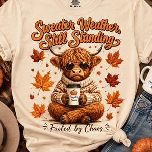 Może przedstawiać: Kremowy t-shirt z grafiką puszystej krowy rasy Highland w okularach przeciwsłonecznych i dzianinowym swetrze. Krowa trzyma kubek kawy z napisem Pumpkin Chaos. Tekst głosi Sweater Weather, Still Standing oraz Fueled by Chaos.