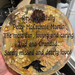 Könnte beinhalten: Ein runder, transparenter Harz-Untersetzer mit getrockneten Blumen und einer schwarzen Vogelsilhouette. Der Text lautet "Rest Peacefully Peter McKechnie Martin The most fun, loving and caring Dad and Grandad. Sorely missed and dearly loved."