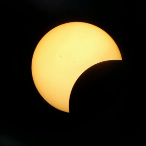 Könnte beinhalten: Foto einer partiellen Sonnenfinsternis. Die Sonne ist eine leuchtend gelb-orangefarbene Sichel vor schwarzem Hintergrund. Mehrere kleine dunkle Flecken sind auf der Sonnenoberfläche sichtbar.
