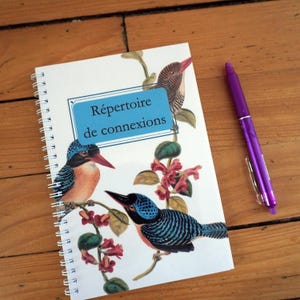 Op de afbeelding: Een spiraalgebonden notitieboek met een omslag met illustraties van kleurrijke vogels en bloemelementen. Het notitieboek heeft de tekst "Répertoire de connexions" op een blauwe achtergrond. Een paarse pen ligt naast het notitieboek op een houten oppervlak.
