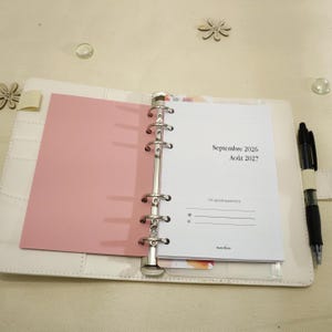 Peut inclure: Un agenda à six anneaux blanc avec un intercalaire rose et des pages blanches. La page ouverte affiche le texte Septembre 2026, Août 2027 et Cet agenda appartient à. Un stylo noir est inséré dans une boucle sur le bord droit de la couverture.