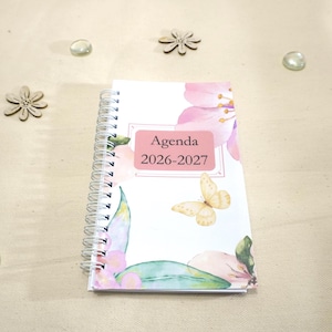 Peut inclure: Un agenda à reliure spirale avec une couverture blanche ornée d'illustrations à l'aquarelle de fleurs roses, de feuilles vertes et d'un papillon jaune. Une étiquette rectangulaire rose affiche le texte noir Agenda 2026-2027.