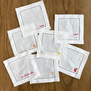 May include: A collection of white linen cocktail napkins with black stitched borders. Some napkins feature embroidered designs of fruit, a cocktail, and a beach umbrella. One napkin has the word "Ciao" embroidered in red.