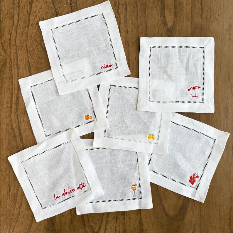 May include: A collection of white linen cocktail napkins with black stitched borders. Some napkins feature embroidered designs of fruit, a cocktail, and a beach umbrella. One napkin has the word "Ciao" embroidered in red.