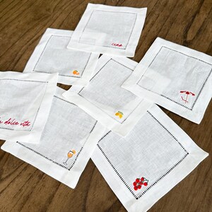 May include: White linen coasters with embroidered details. Some coasters feature the text "Ciao" and "La dolce vita" in red. Other coasters have embroidered images of a cocktail, fruit, and a beach umbrella.